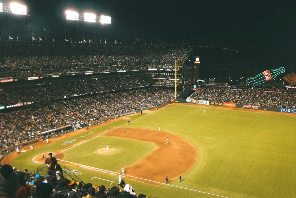 스탠드에서 바라본 붐비는 경기장의 야간 야구 경기로, 경기장에 있는 선수들과 조명이 켜져 있습니다.