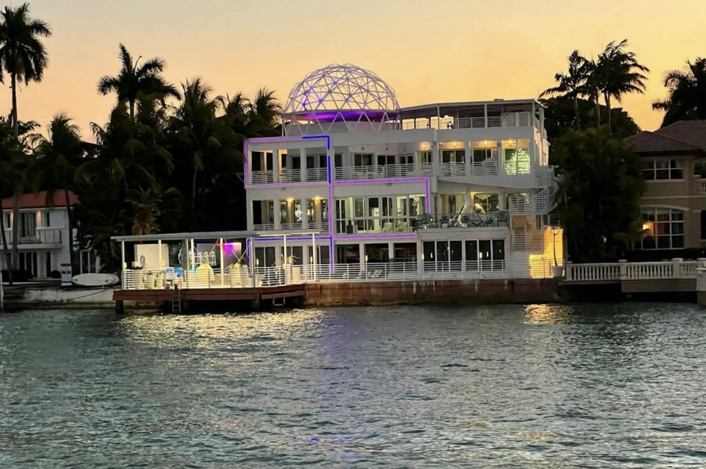 석양에 열대 나무로 둘러싸인 지붕에 조명 돔 구조의 현대적인 대형 수변 주택입니다.