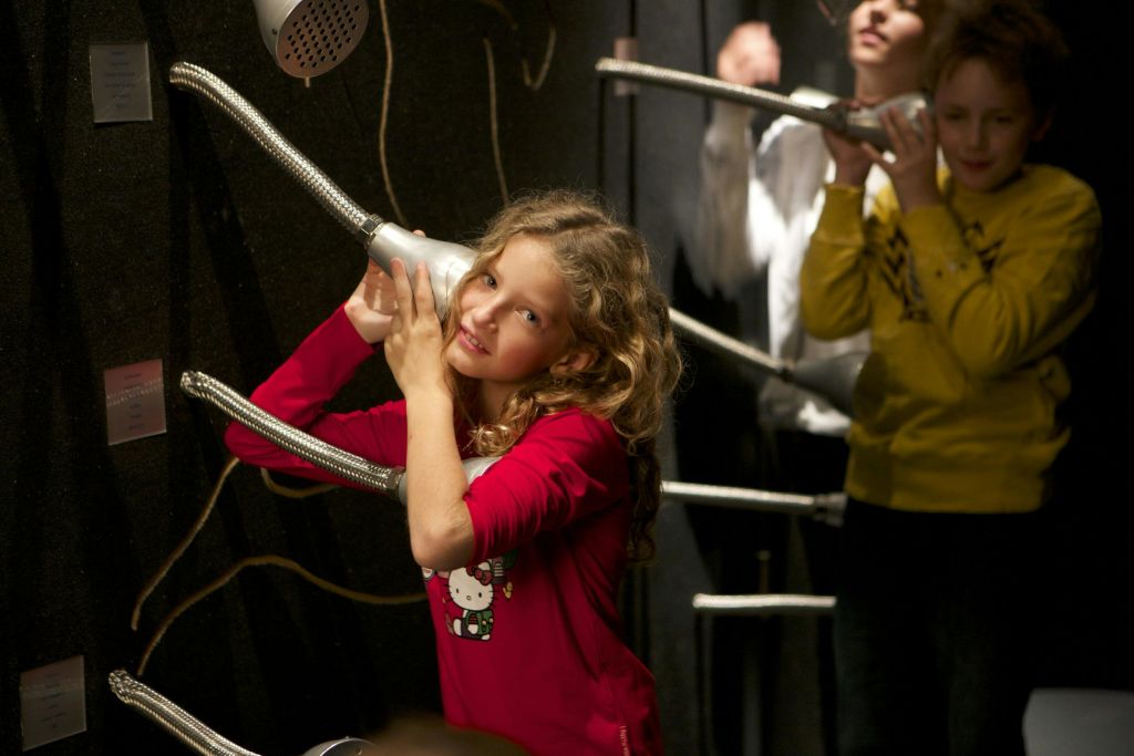 빨간색 셔츠를 입은 아이 3명과 노란색 셔츠를 입은 아이 2명이 튜브 커뮤니케이션 전시물과 상호작용합니다.