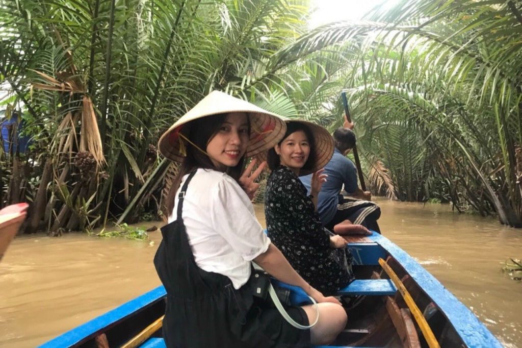 원뿔 모자를 쓴 두 여성이 보트에 앉아 울창하고 좁은 수로를 헤매며 미소 짓고 제스처를 취하고 있습니다.