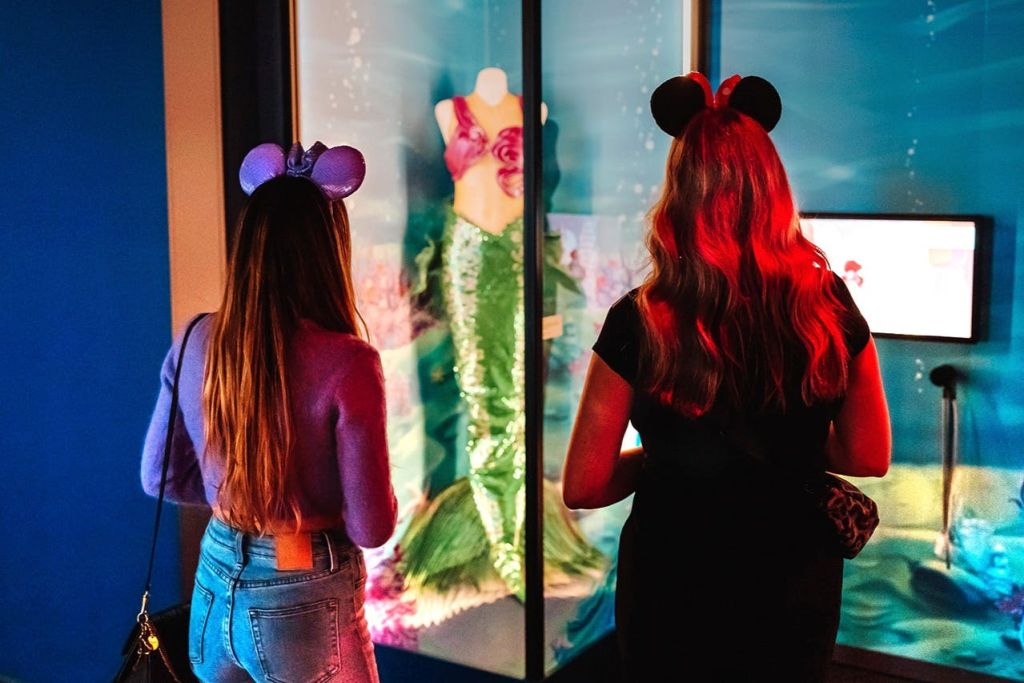 마우스 이어 헤드밴드를 착용한 두 명의 여성이 디스플레이 케이스 앞에 서서 초록색 꼬리와 분홍색 상의가 달린 인어 의상을 선보이고 있습니다.