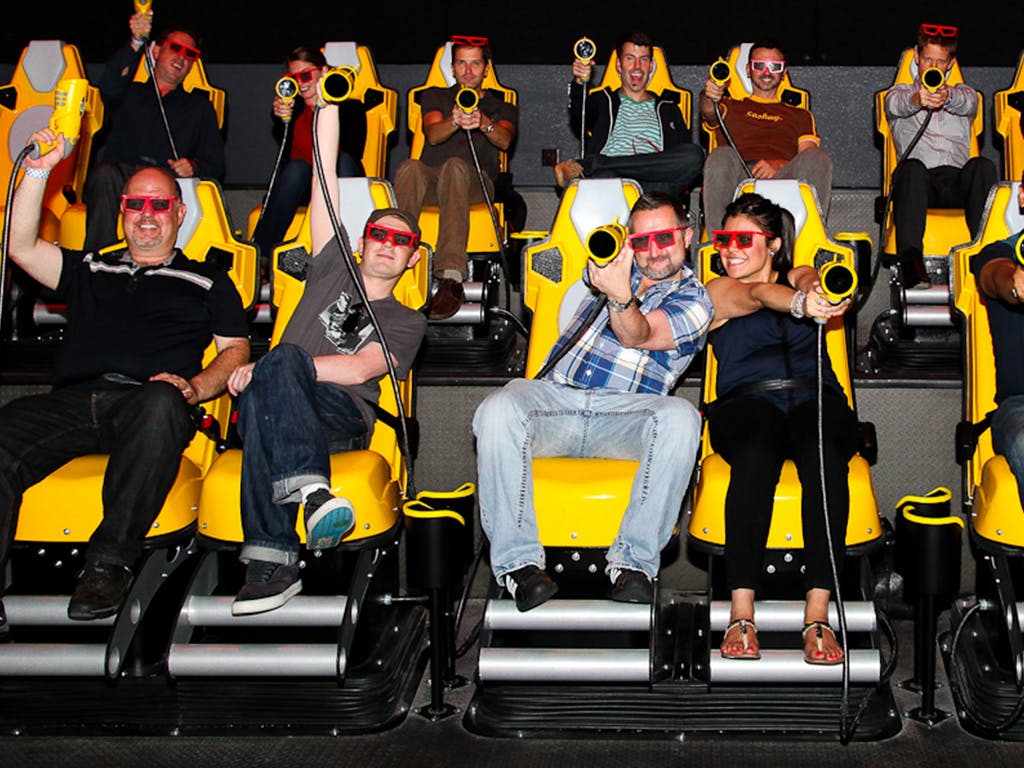 노란색 모션 시뮬레이터 의자에 앉은 3D 안경을 쓰고, 경총을 들고, 웃으며 대화형 라이딩에 참여하는 사람들.