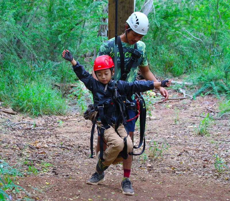 붉은 헬멧을 쓰고 하네스를 착용한 어린이가 두 팔을 뻗은 채 숲 속 하얀 헬멧을 쓴 성인의 도움을 받으며 걷고 있습니다.