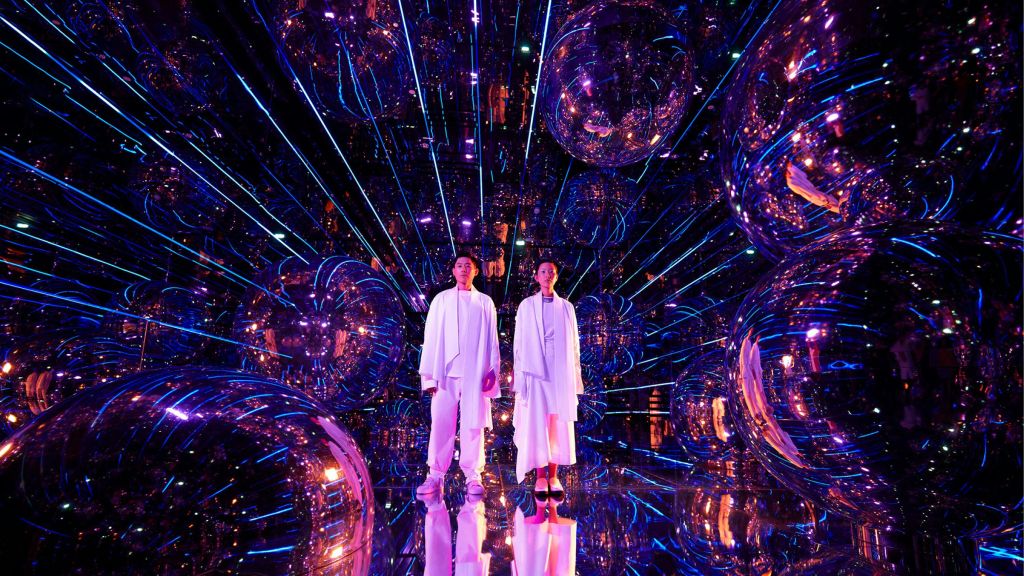 흰색 옷을 입은 두 사람이 반사 표면, 커다란 거울 구체, 선명한 보라색과 파란색 조명으로 가득한 방에 서 있습니다.