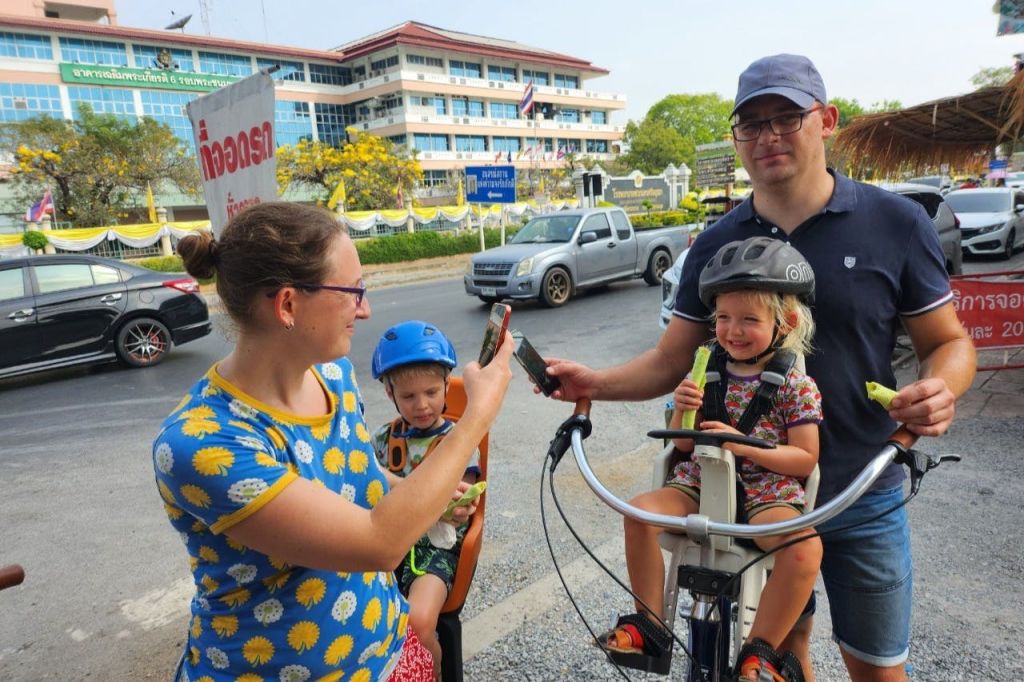어린이 자전거 좌석에 한 명씩, 건물이 배경으로 있는 도로 근처의 야외에서 두 자녀를 둔 가족.