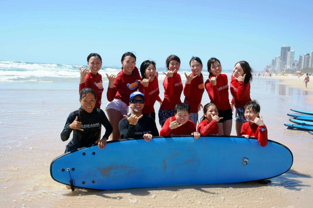 빨간 셔츠를 입은 한 무리의 사람들이 해변에서 서핑 보드를 들고 포즈를 취하며 샤카 간판을 만들고 미소를 짓고 있습니다. 파도는 배경에서 볼 수 있습니다.