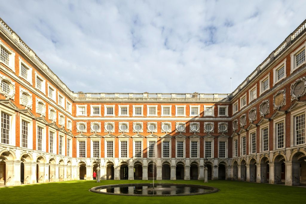 둥근 중앙 분수가 있는 대칭형 안뜰로, 부분적으로 흐린 하늘 아래 아치형 창문이 있는 붉은 벽돌 건물로 둘러싸여 있습니다.