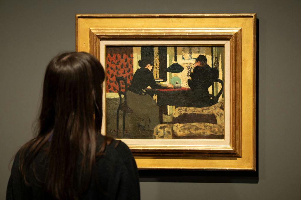 한 사람이 박물관에서 액자 그림을 관찰하고 있는데, 국내 인테리어의 테이블에 앉아 있는 두 사람의 모습이 그려져 있습니다.