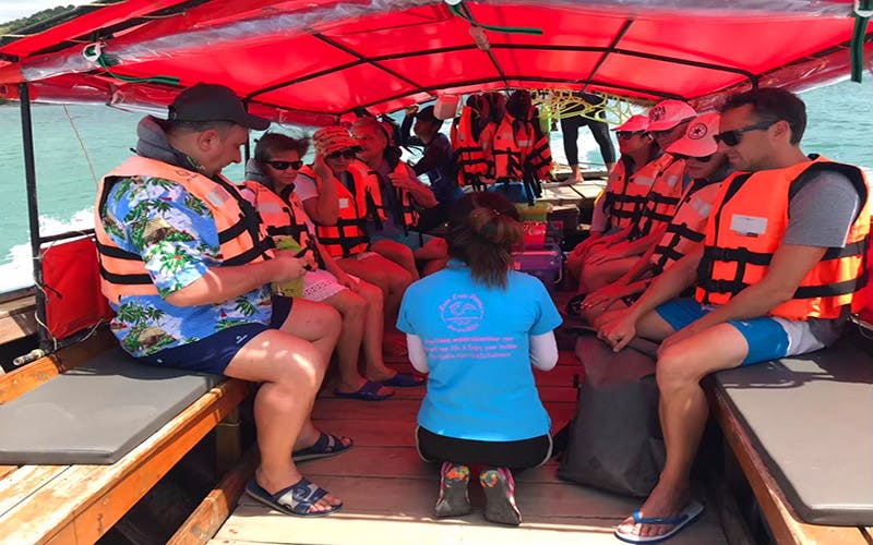 구명조끼를 입고 빨간색 캐노피 아래 보트에 앉아 있는 사람들. 파란색 셔츠를 입은 한 사람이 무릎을 꿇고 있습니다.