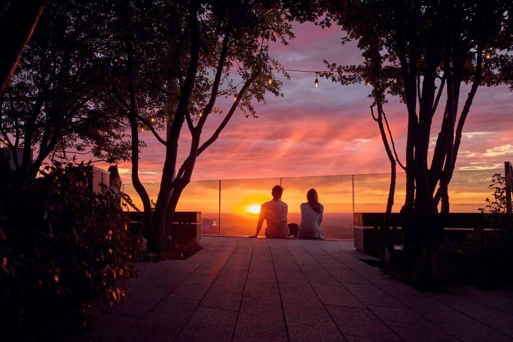 두 사람이 옥상 바닥에 앉아 나무와 줄 조명으로 둘러싸인 유리 장벽을 통해 생생한 석양을 감상하고 있습니다.