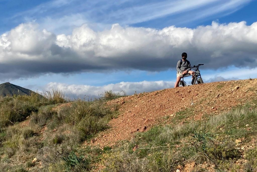 회색 후드티를 입은 사람이 마른 잔디와 부분적으로 흐린 하늘을 배경으로 언덕 꼭대기에서 자전거를 타고 휴식을 취하고 있습니다.