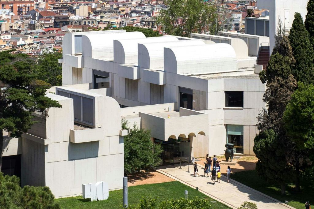 녹음과 통로로 둘러싸인 곡선형 지붕이 돋보이는 현대적인 흰색 건물로, 도시 풍경을 배경으로 한 건물입니다.