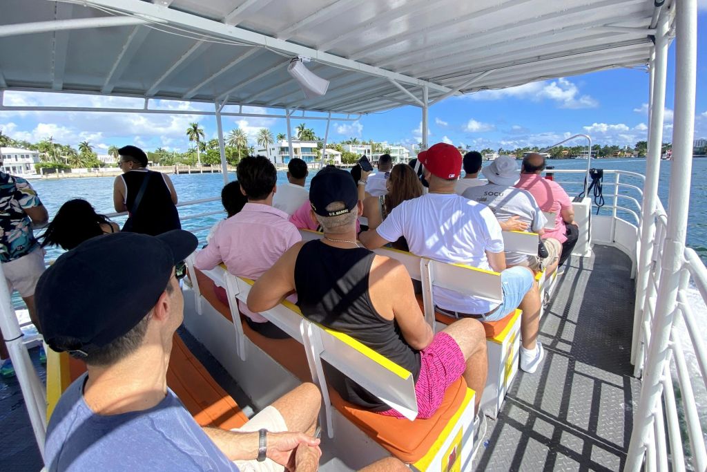 사람들은 지붕이 덮인 보트에 앉아 정면을 향하고 있습니다. 푸른 하늘 아래 물, 야자수, 하얀 건물이 배경으로 보입니다.