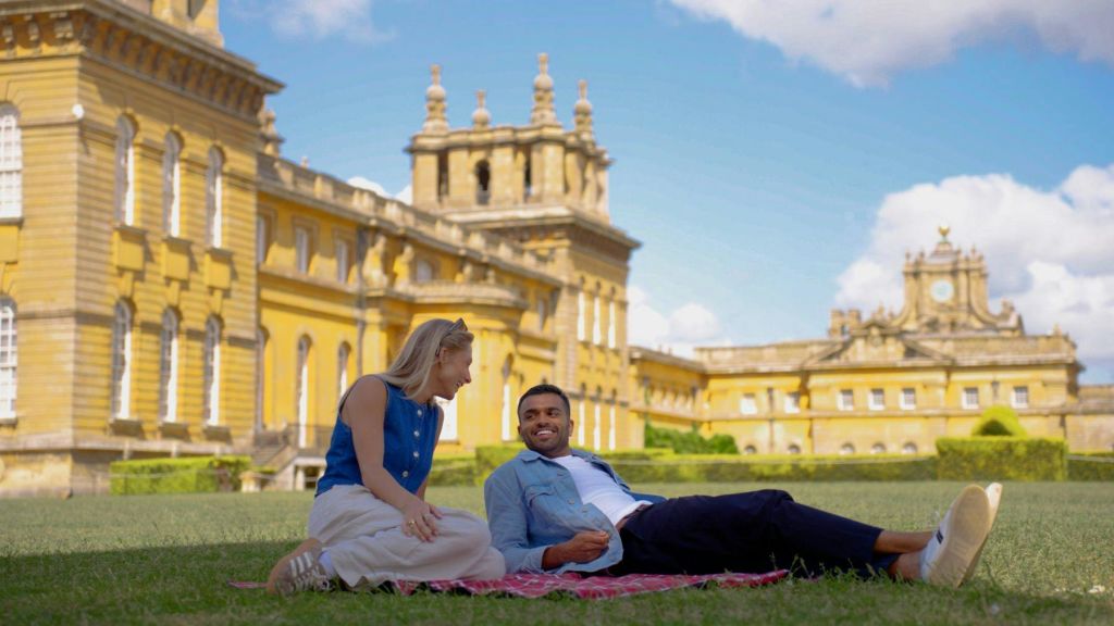 녹색 잔디밭의 빨간색 피크닉 담요 위에 앉아 타워가 있는 유서 깊은 노란색 건물 앞에서 서로 미소 짓고 있습니다.