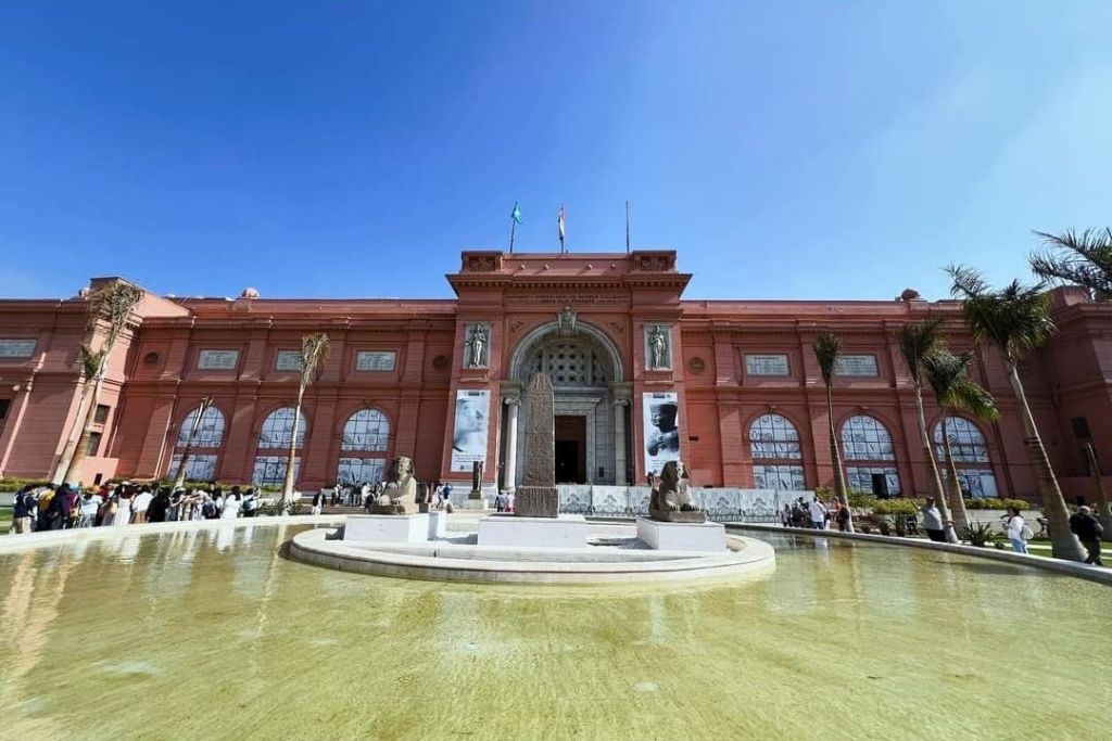 직사각형 수영장 앞에 중앙 오벨리스크를 둘러싼 아치형 입구, 기둥, 조각상이 있는 빨간색 건물입니다. 위쪽의 맑고 푸른 하늘.