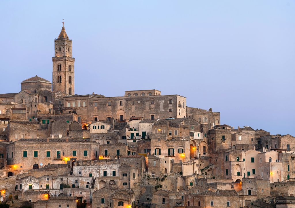 촘촘하게 들어선 석조 건물과 맑은 저녁 하늘 아래 높이 솟은 종탑이 있는 오래된 언덕 마을입니다.