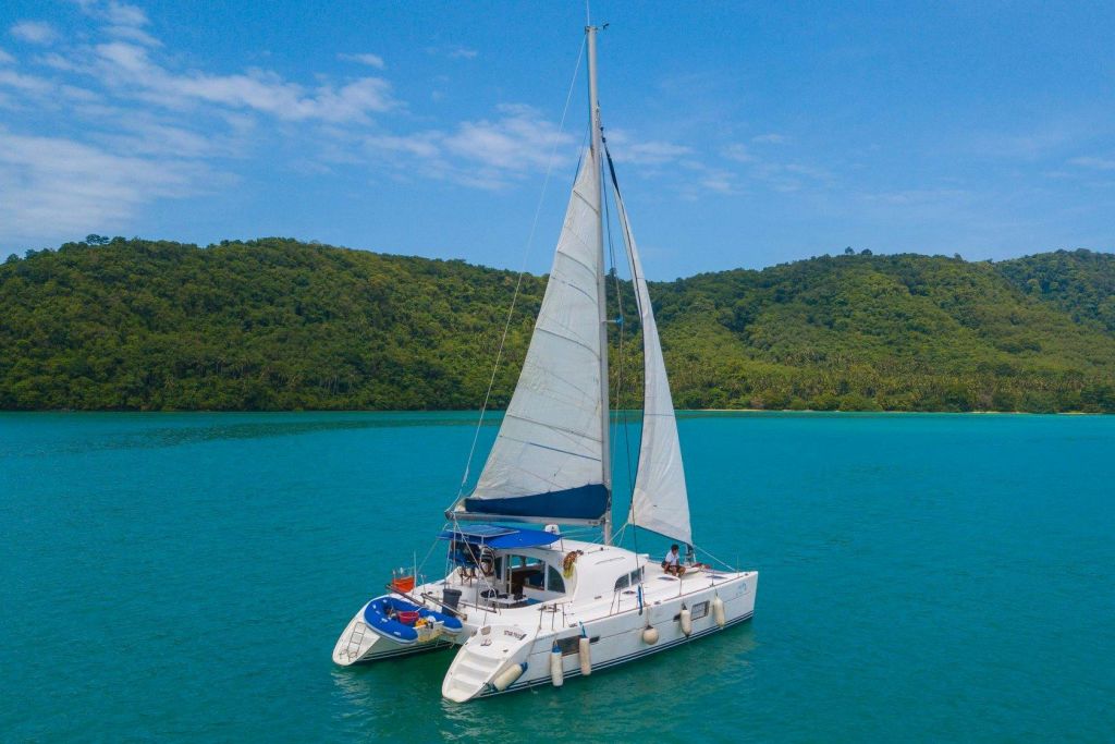 돛을 올린 흰 쌍동선이 부분적으로 흐린 하늘 아래 푸르고 숲이 우거진 해안가 근처의 청록색 물가에 정박해 있습니다.