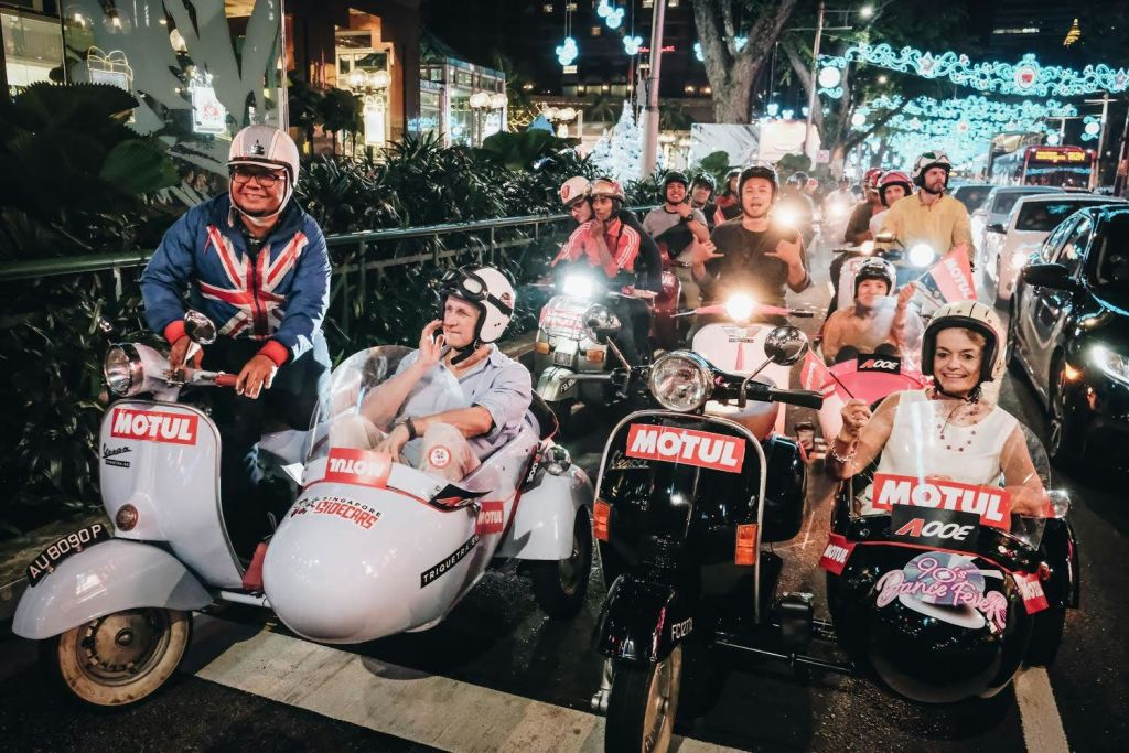 한 무리의 사람들이 밤에 오토바이와 사이드카를 타고 도시의 거리를 내려가며 손을 흔들고 미소를 짓고 있습니다.