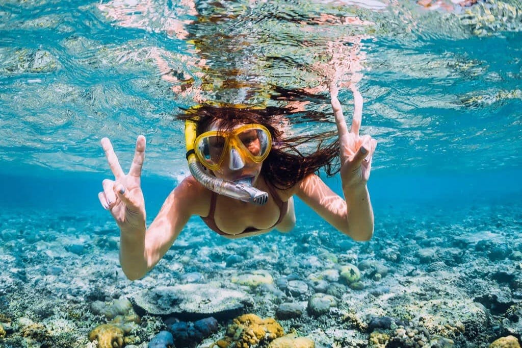 수중에서 스노클링과 마스크를 착용하고 산호와 맑고 푸른 물로 둘러싸인 두 손으로 평화 표지판을 들고 있는 사람.