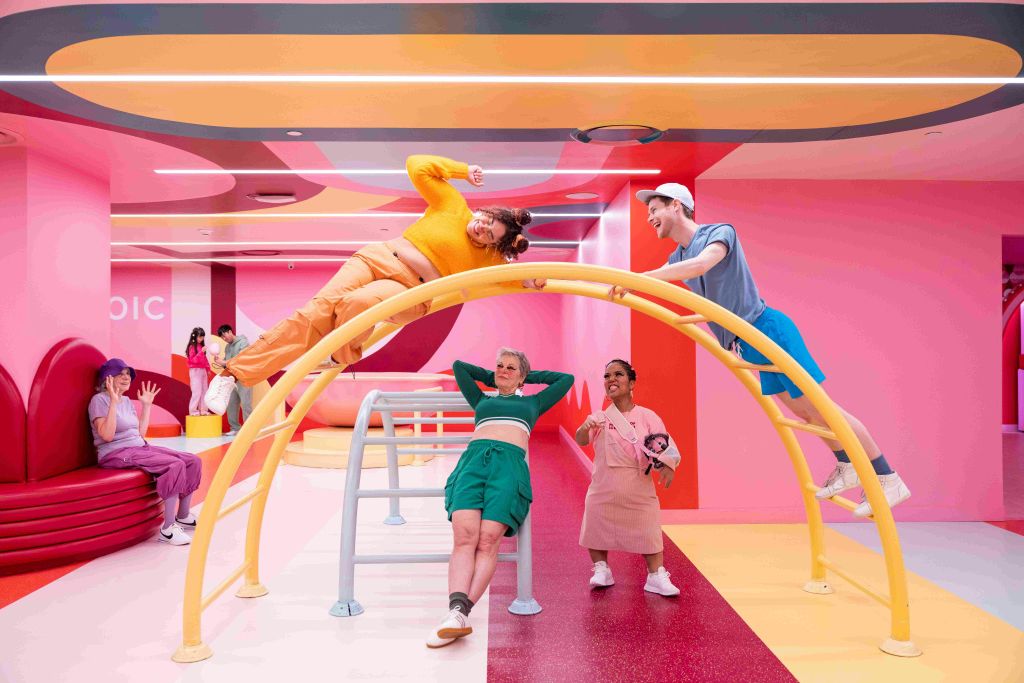 다채로운 색상의 의상을 입은 성인 4명이 활기찬 파스텔 색상의 실내 공간에서 놀이터 장비와 상호 작용합니다.