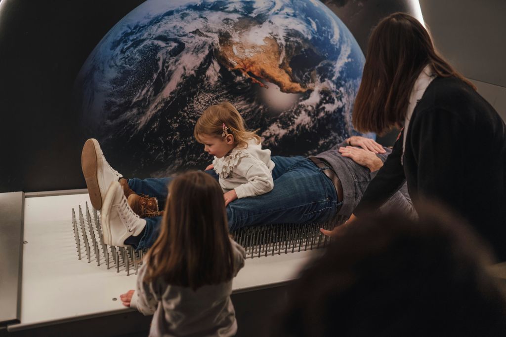 한 어린이가 손톱 침대에 누워 있는 어른 위에 앉아 있는데, 지구가 배경으로 표시됩니다. 한 소녀가 근처를 지켜보고 있습니다.