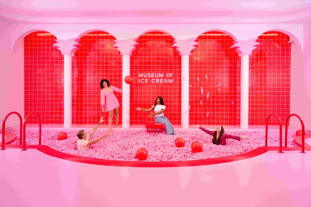 아이스크림 박물관의 분홍색 물체가 가득한 대형 수영장에서 4명이 놀고 있습니다. 빨간색 타일과 흰색 기둥이 배경에 있습니다.