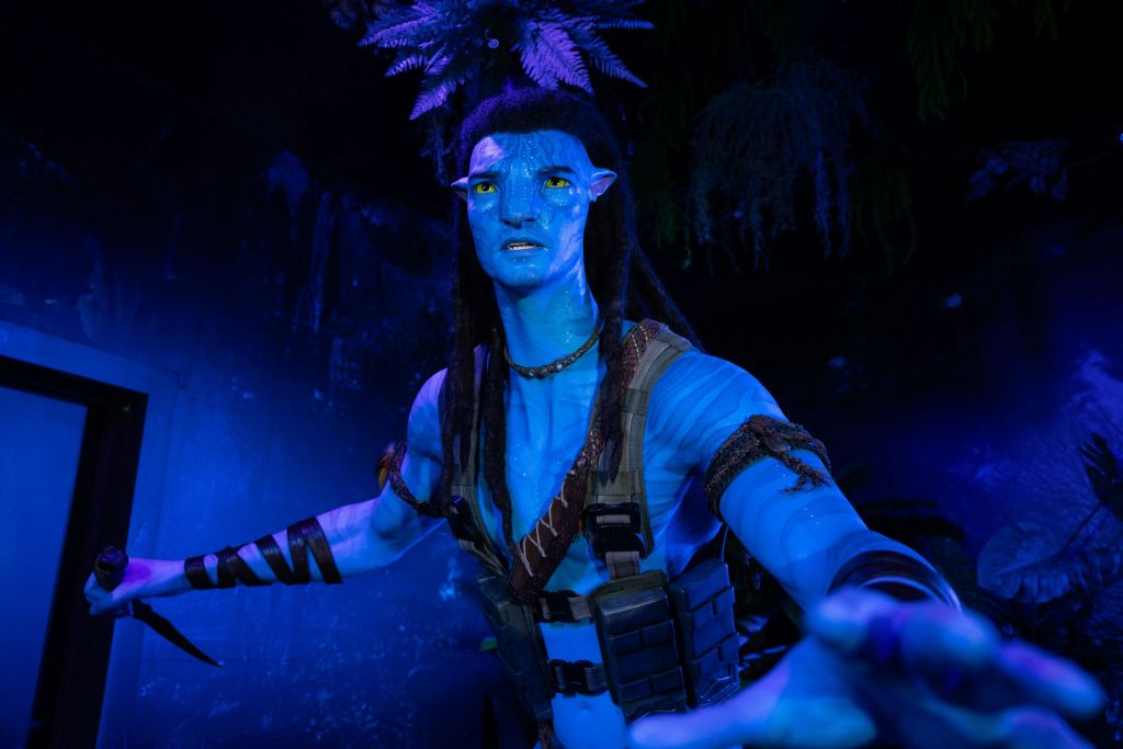 희미한 조명 환경에서 부족적이고 전술적인 복장을 하고 빛나는 노란 눈과 땋은 머리를 가진 푸른 피부의 휴머노이드 피규어.