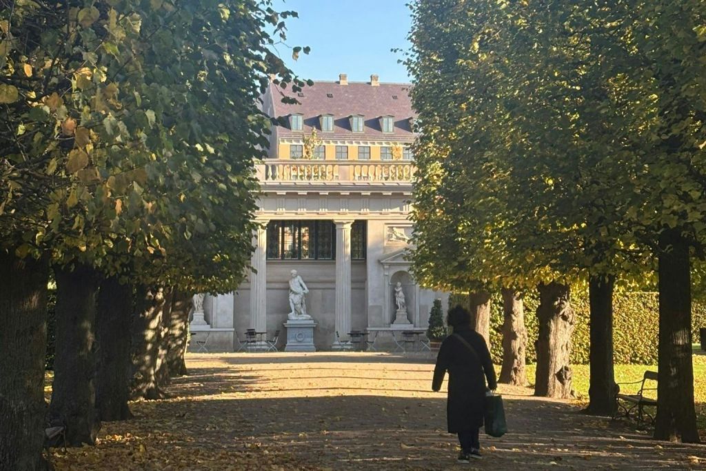 화창한 날에 조각상과 기둥이 있는 웅장한 건물을 향해 나무가 늘어선 길을 걷는 사람.