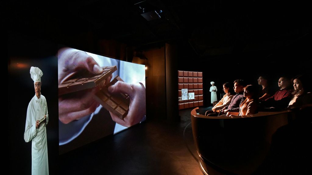 어두운 방에 앉아 초콜릿 바를 깨는 손을 보여주는 대형 스크린을 보고 있는 사람들, 근처에 조명 마네킹이 서 있습니다.