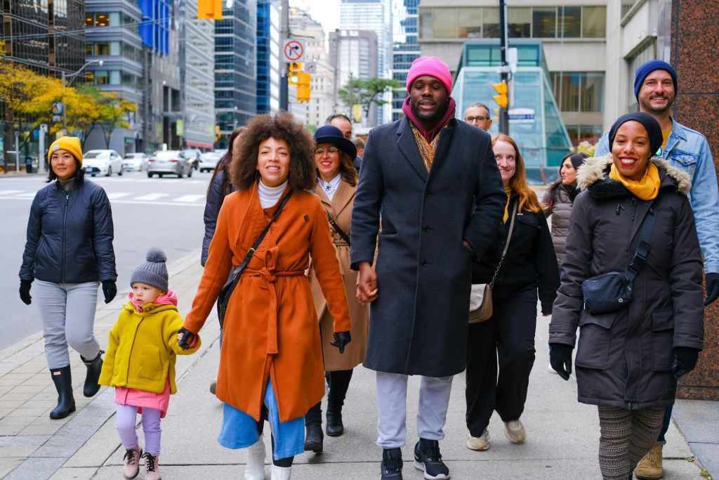 밝은 날 한 무리의 사람들이 도시 보도를 걷고 있습니다. 중심부 사람들은 화려한 색의 코트를 입고 있습니다.