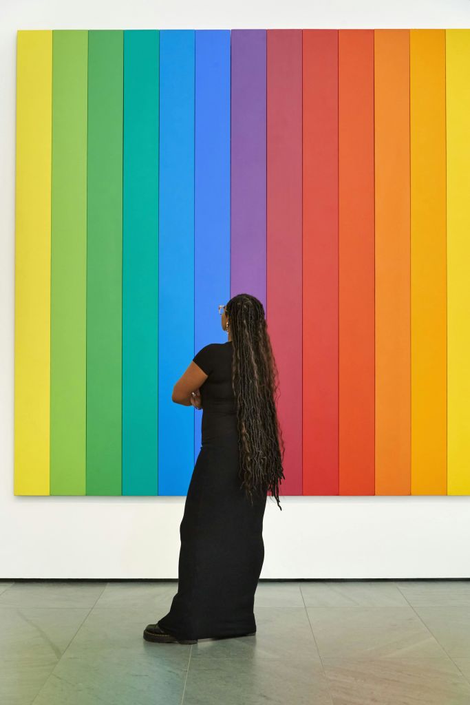 흰 벽에 그려진 커다란 무지개색 줄무늬 작품을 바라보는 긴 머리의 사람.