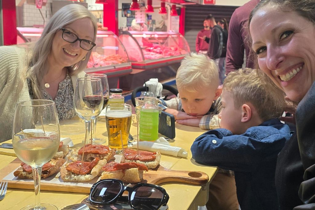 고기 카운터를 배경으로 두 명의 여성과 두 명의 어린이가 시장의 테이블에 앉아 음료와 음식을 즐기고 있습니다.