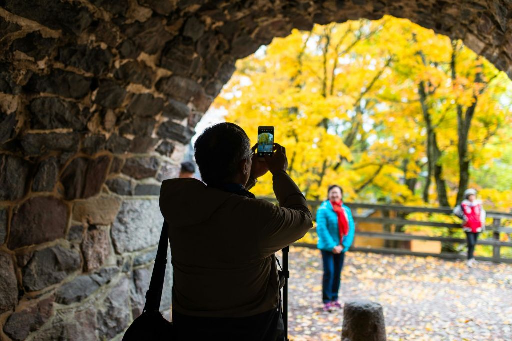 파란색 재킷과 빨간색 스카프를 입은 사람이 가을 나무를 배경으로 돌 아치 프레임으로 사진을 찍고 있습니다.