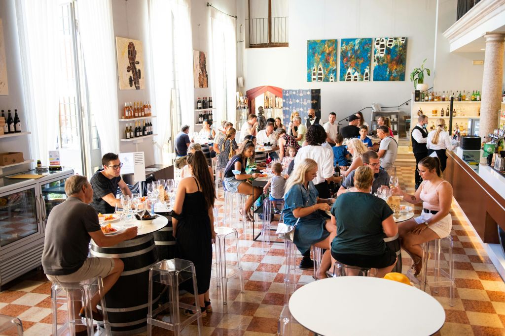 높은 창문, 선반에 놓인 와인병, 벽에 걸린 그림이 있는 밝은 레스토랑에서 식사를 하고 사람들과 어울립니다.