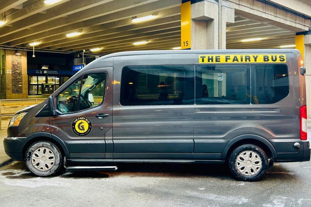 측면에 "The Fairy" 와 로고가 있는 회색 셔틀 밴이 "15" 라고 표시된 노란색 기둥 옆에 콘크리트 구조로 주차되어 있습니다.