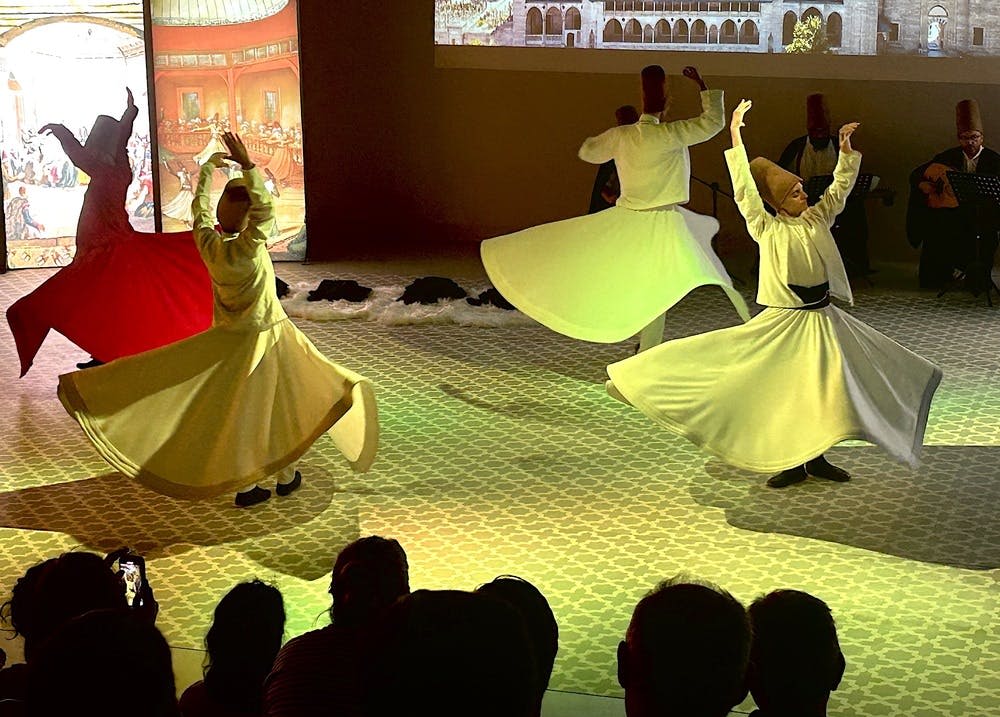 흰 가운을 입은 소용돌이는 무늬가 있는 바닥에서 춤을 추고, 청중은 전경에 앉아 춤을 춘다.