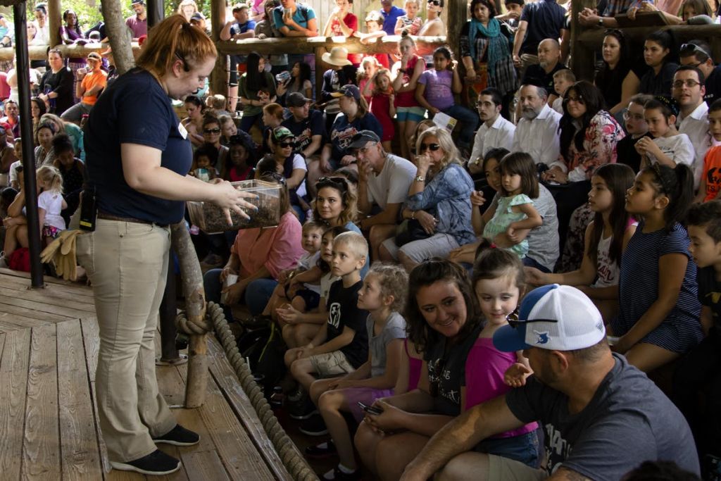 발표자가 야외에서 자리에 앉은 청중에게 아이템을 보여줍니다. 군중에는 어른과 아이들이 세심하게 지켜보고 있습니다.