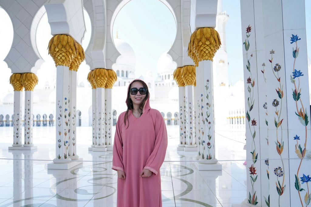 분홍색 가운과 선글라스를 입은 여성이 장식 기둥과 황금색 디테일이 있는 밝고 화려한 안뜰에 서 있습니다.