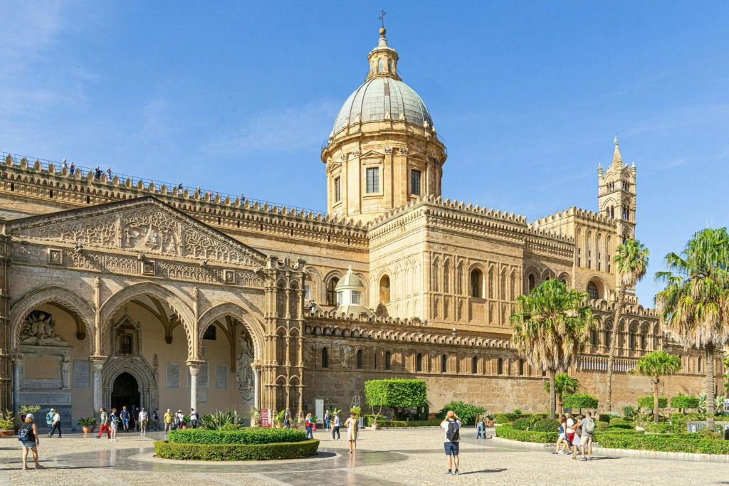 정교한 건축물과 맑고 푸른 하늘 아래 커다란 돔이 있는 유서 깊은 대성당으로 관광객들이 아래 광장을 둘러보고 있습니다.