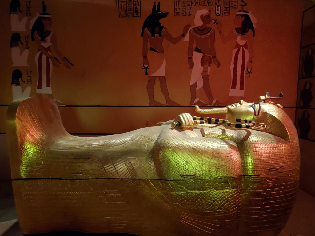 아누비스와 다른 신들을 포함한 상형문자와 인물이 그려진 이집트 벽화 앞에 있는 황금 석관.