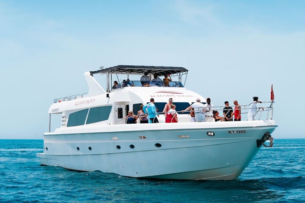 맑고 푸른 하늘 아래 갑판에 여러 사람이 있는 탁 트인 바다 위의 하얀 요트.