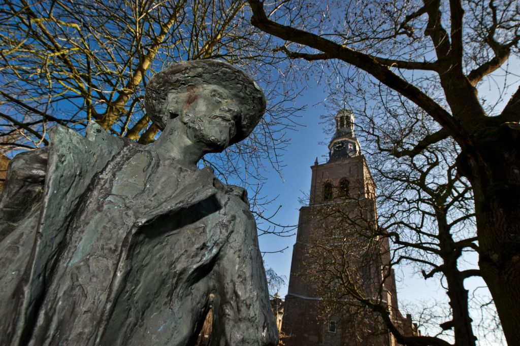 근처에 시계탑이 있고 푸른 하늘을 배경으로 잎이 없는 나무가 있는 모자를 쓴 남자의 청동 동상.