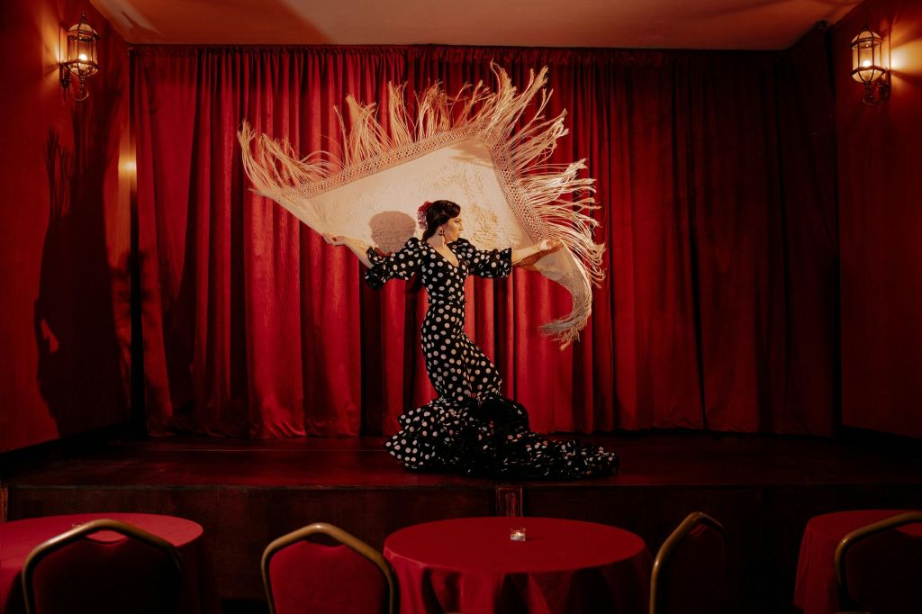 검은색과 흰색 물방울무늬 드레스를 입은 댄서가 무대 전면에 빨간색 커튼과 의자가 있는 프린지 숄을 들고 있습니다.