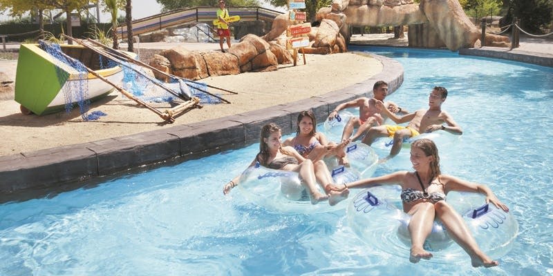 워터 파크의 느긋한 강에서 풍선 튜브를 타고 수영복을 입은 네 명이 구조대와 표지판을 배경으로 휴식을 취하고 있습니다.