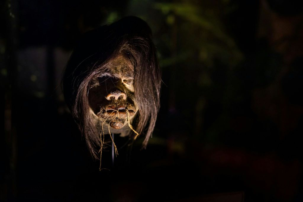 길고 어두운 머리카락과 실이 감긴 입술과 눈을 가진 유령처럼 디테일한 쪼그라든 머리를 보여주는 희미한 조명 장면.