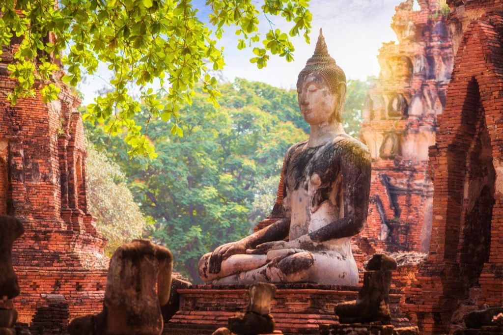 부드러운 햇빛 아래 고대 벽돌 구조물과 나무들 속에서 명상을 하며 앉아 있는 풍화된 불상입니다.