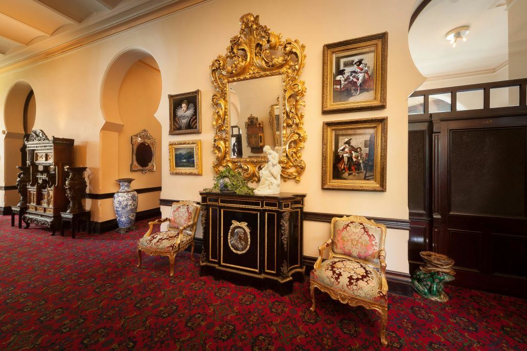 골드 프레임 거울, 액자 페인팅, 벨벳 덮개 의자, 검은색 수납장, 레드 카펫의 장식 화병이 있는 화려한 객실입니다.