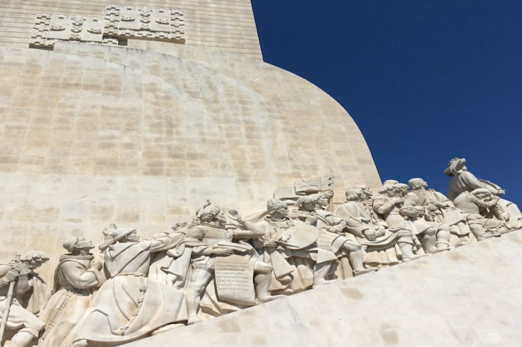 커다란 돌담과 깊고 푸른 하늘을 배경으로 디테일한 로브를 입은 역사적인 인물들의 행렬이 특징인 석조 조각.