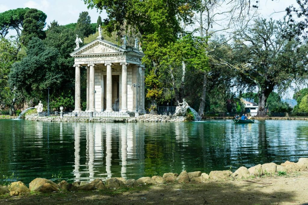 나무로 둘러싸인 연못 옆의 신고전주의 양식의 사원으로, 물 위에 작은 배가 보입니다.