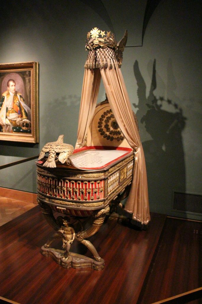 박물관 벽에 걸린 역사적인 유니폼을 입은 남자의 초상화 옆에 화려한 디테일과 장식된 직물로 장식된 왕실 요람.